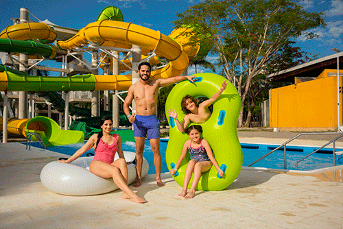 Familia de cuatro personas, mujer, hombre, un niño y una niña posando con flotadores en forma de ocho para dos personas, uno blanco y uno verde junto a un tobogán amarillo con verde, que finaliza en la piscina que esta detrás de la familia la cual disfruta el día soleado y los descuentos en Lagosol y Lagomar